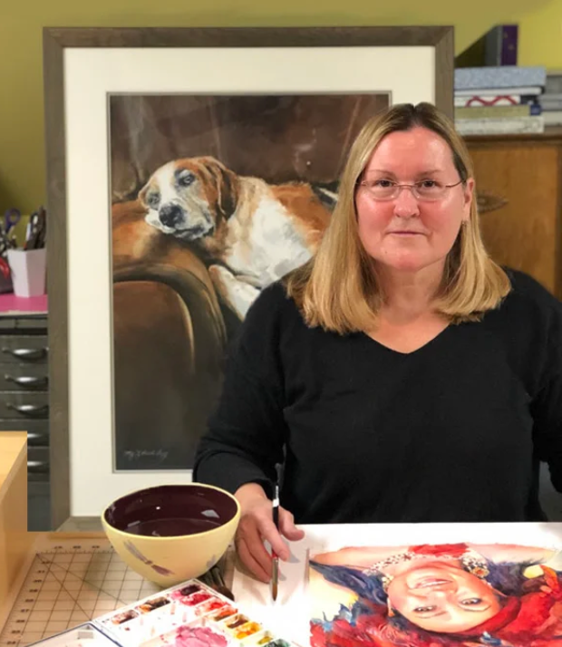 Image of Artist Stacy Lund Levy sitting at her art table painting a portrait with a Beagle on a coach painting in the background.